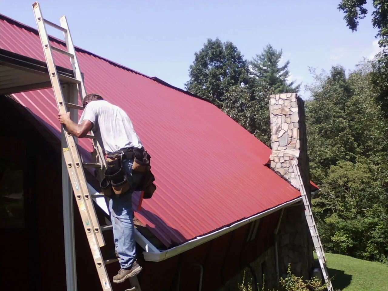Roofer on a ladder beside a red metal roof and stone chimney — Soffit & Fascia Repair by Roofing Brothers in Mohegan Lake