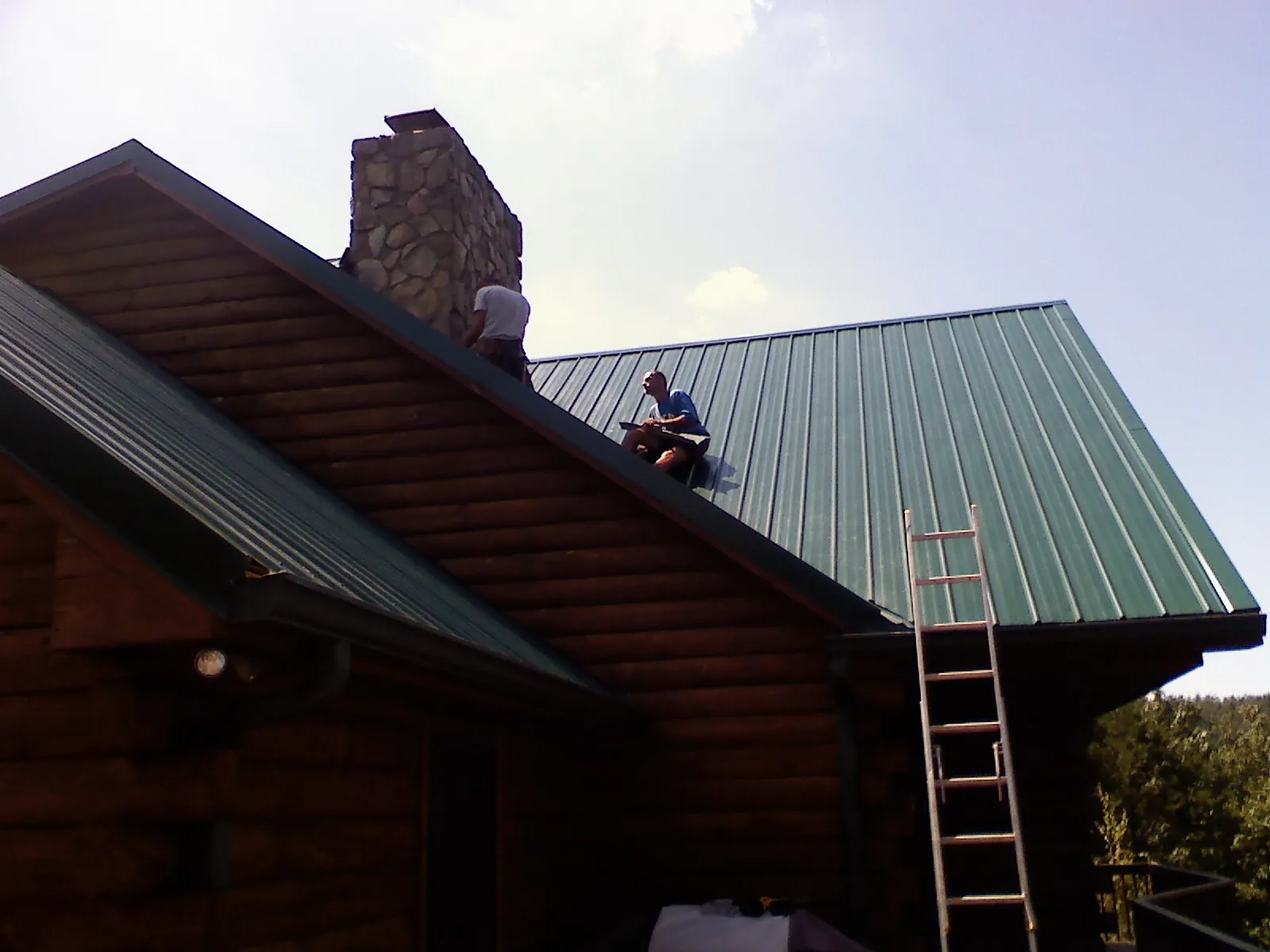 Roofers installing green metal panels beside a stone chimney on a log home in Mohegan Lake